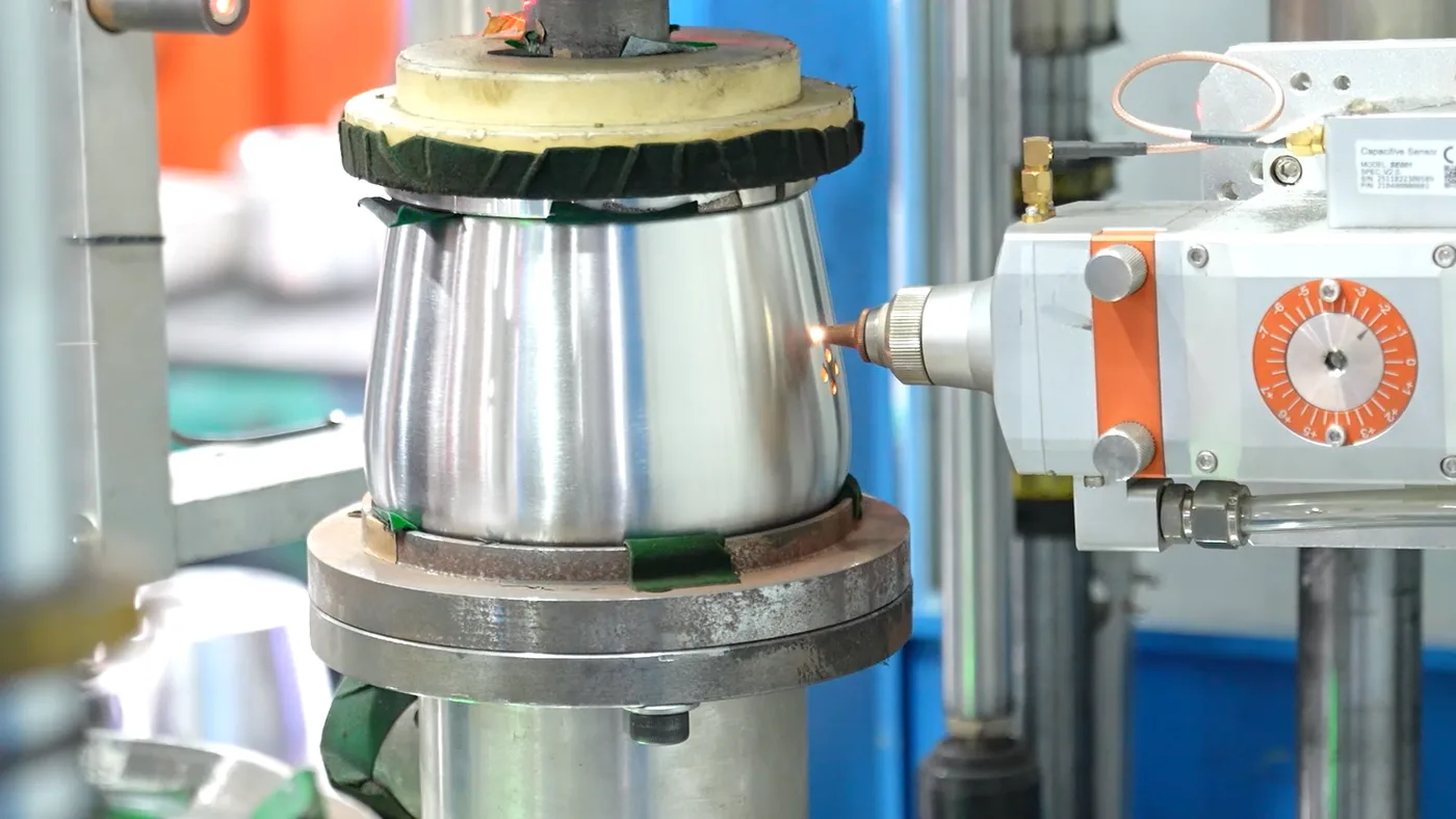 A stainless steel kettle body clamped in a rotating fixture while a torch flame heats the joint on a brazing station